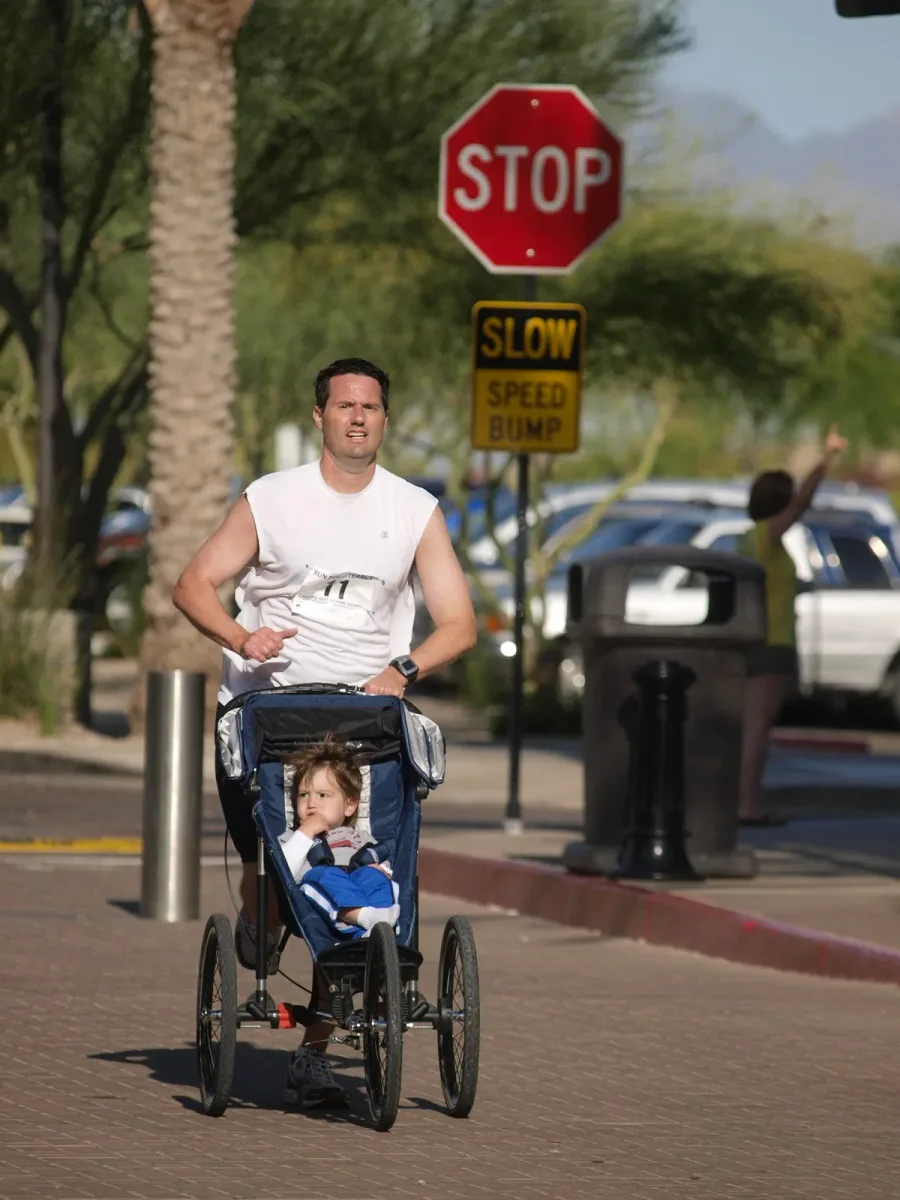 Patrick running a 5K in a sleeveless shirt, pushing a toddler in a jogging stroller