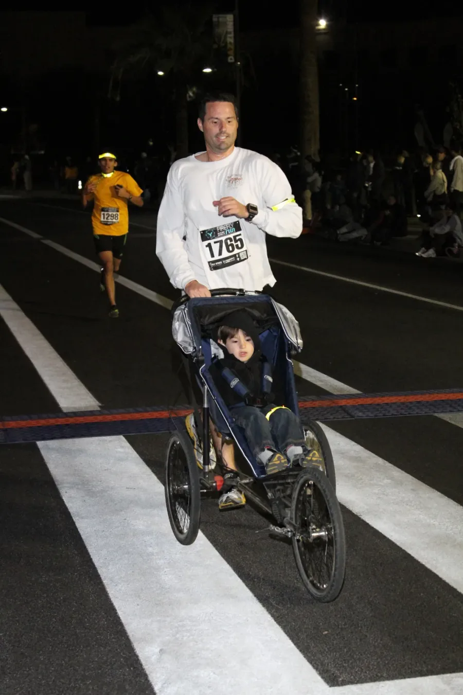 Patrick crossing the finish line of a nighttime race, pushing a jogging stroller with a bundled-up little boy inside