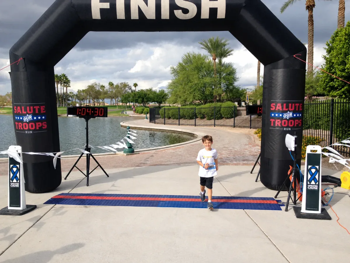 A young boy smiling as he crosses the finish line of a 5K, with the race clock reading 1:04:30