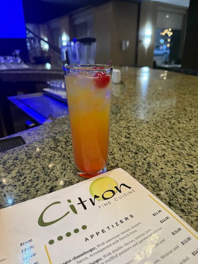 A tall orange cocktail with a cherry on a bar, in front of a Citron Fine Cuisine menu