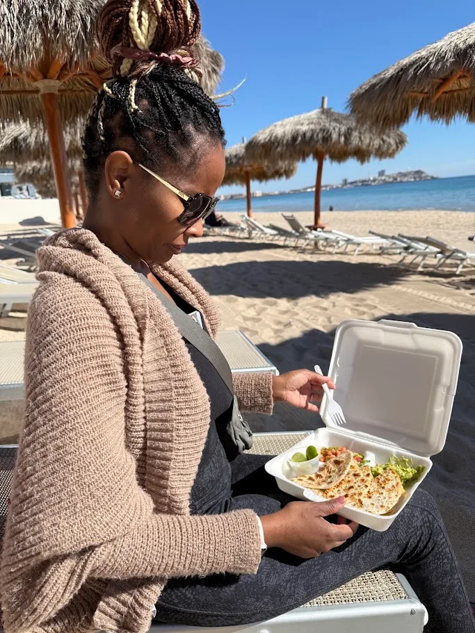 Tamekia eating a to-go lunch on a beach chair under palapa umbrellas, with the water behind her