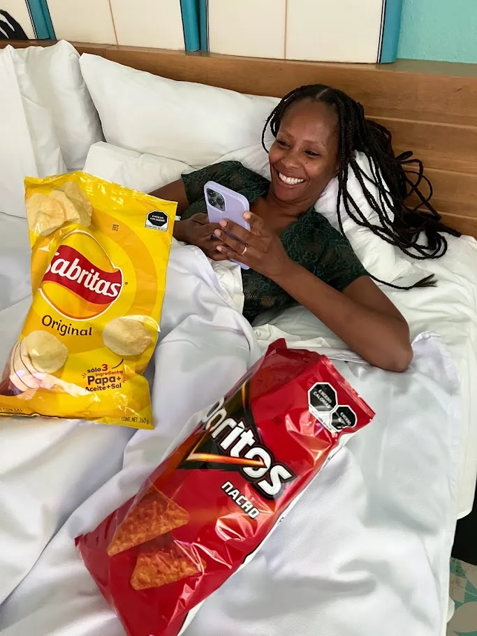 Tamekia smiling at her phone in bed with a big bag of Sabritas and a bag of Doritos Nacho